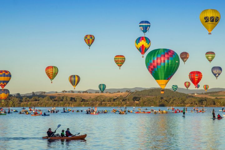 Hot air balloons in sky over lake with kayakers below.