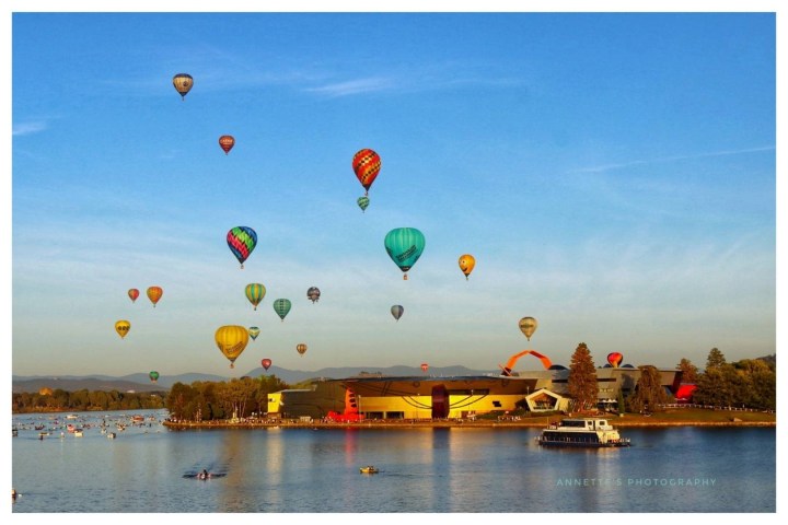 Multiple colorful hot air balloons in the sky above a river and a building during daytime.