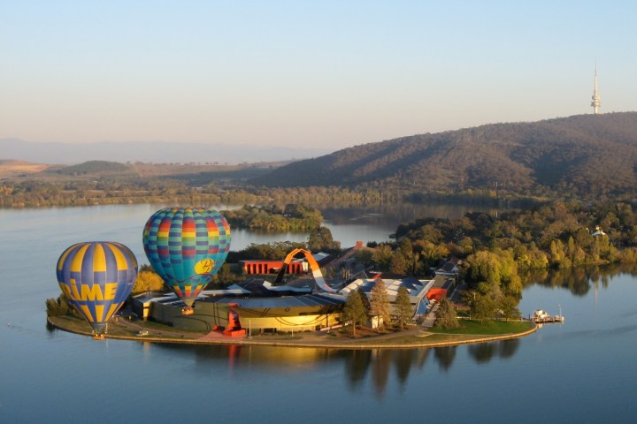Hot air balloons floating over a lake with a hilly landscape and tower in the background.