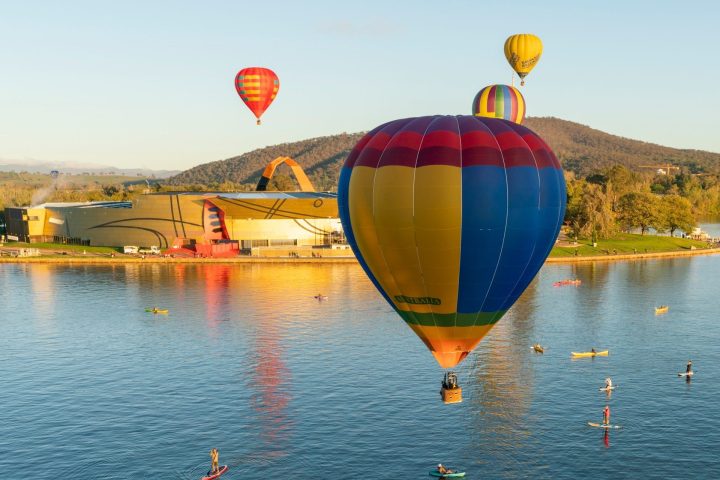 Colorful hot air balloons over a calm lake with kayakers below.