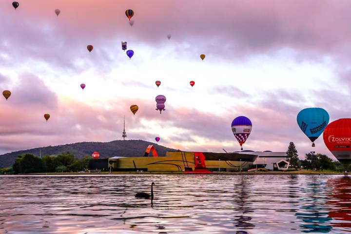 Hot air balloons floating over a lake with hills and buildings in the background at sunrise.