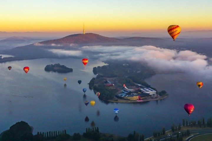 Multiple hot air balloons over lake at sunrise, near a hill with a tower, and surrounded by mist.